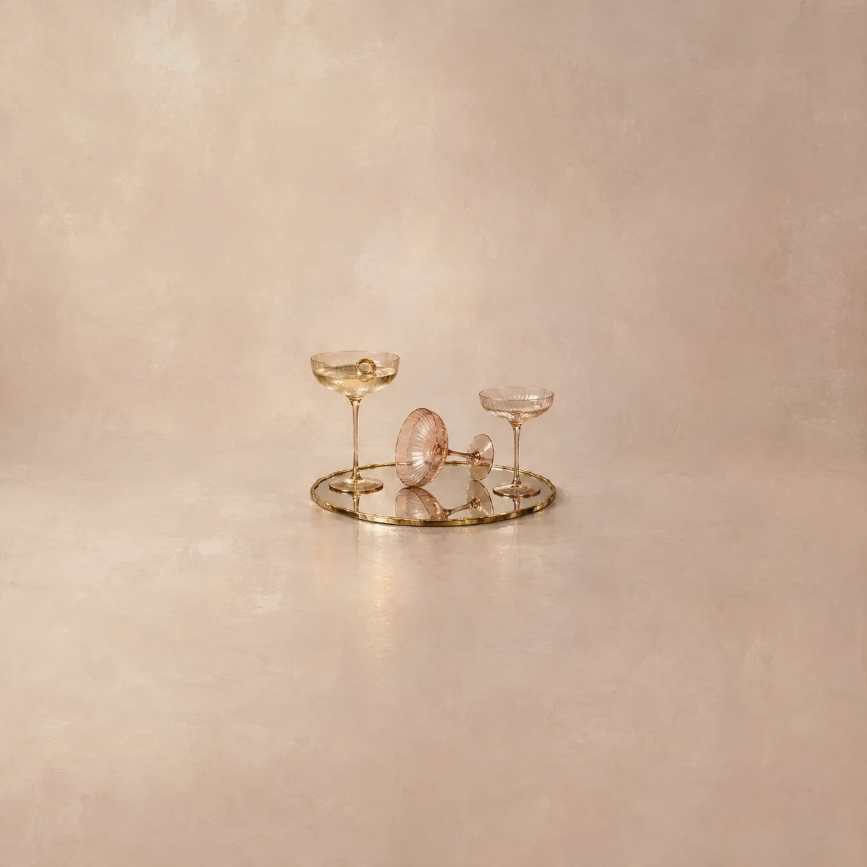 Three stemmed glasses, one tipped sideways, rest on a round gold-edged mirror against pale peach.