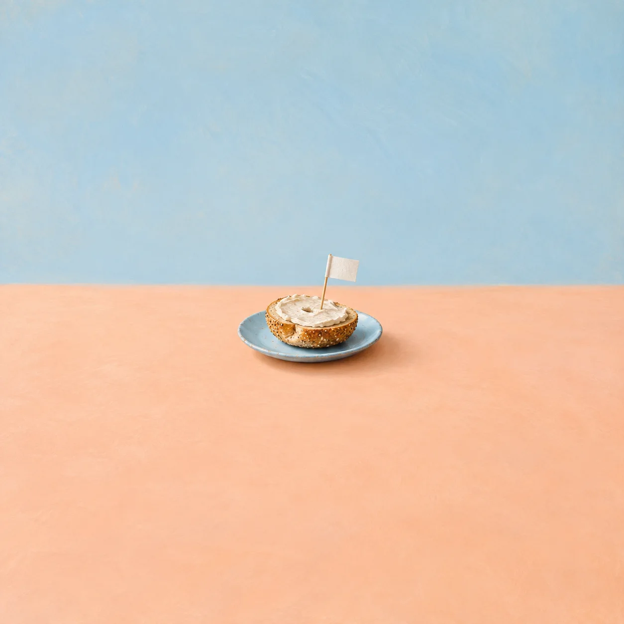A sliced bagel with pale spread and a small blank flag on a blue plate against peach and sky-blue surroundings.