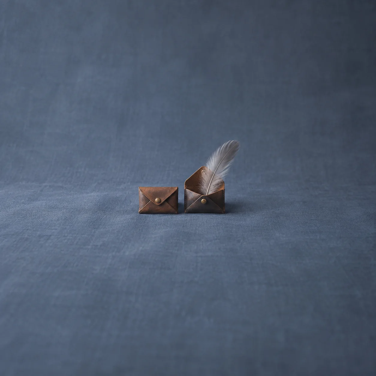 A closed leather pouch beside an open pouch holding a grey feather on blue cloth.