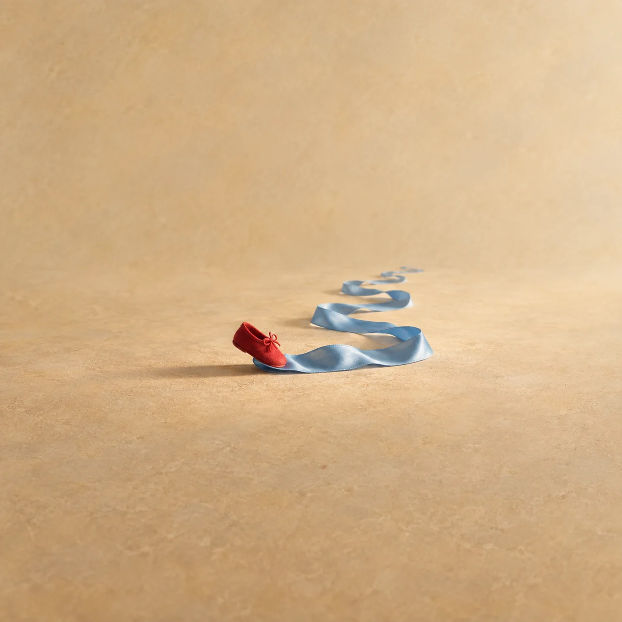 A blue silk ribbon winding across a sandy surface from a small red slipper.