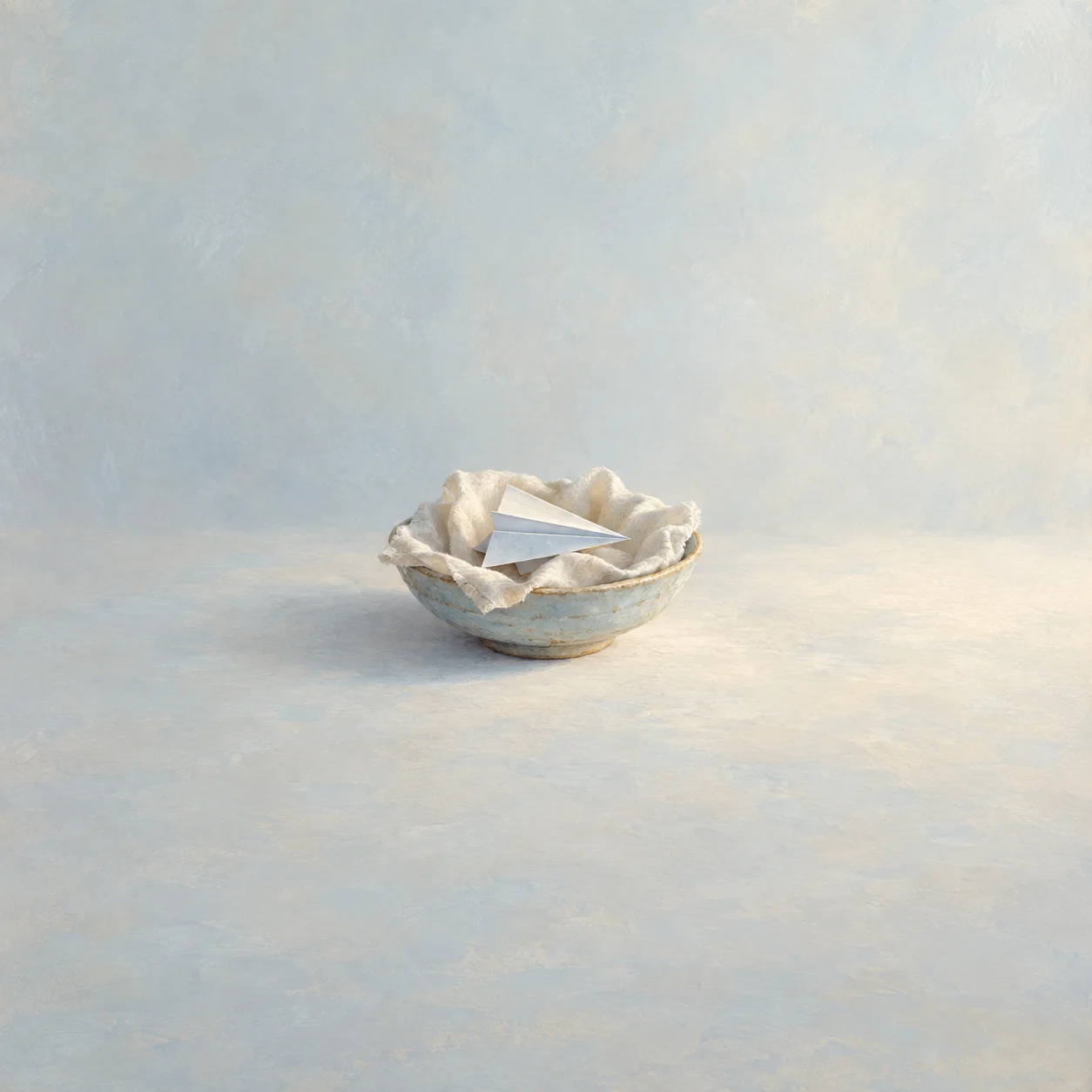 A folded white paper airplane rests in a cloth-lined shallow bowl against a pale blue and cream background.