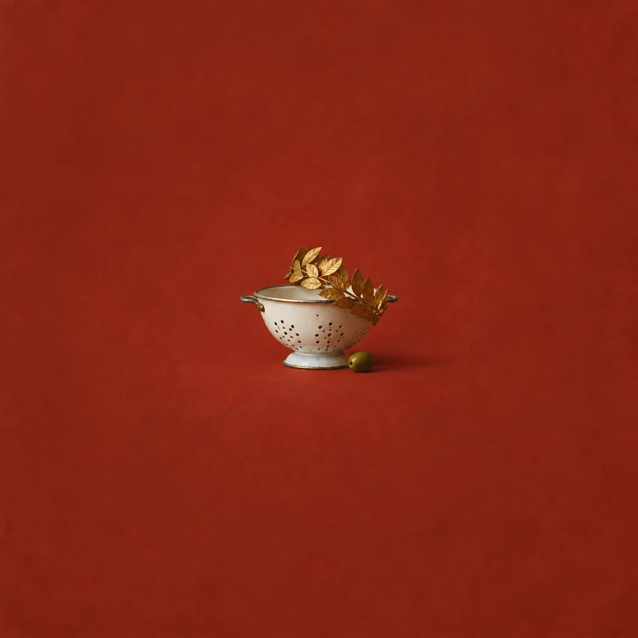 A small gilded laurel crown tilted on a white enamel colander with a green olive beside it against a brick-red background.
