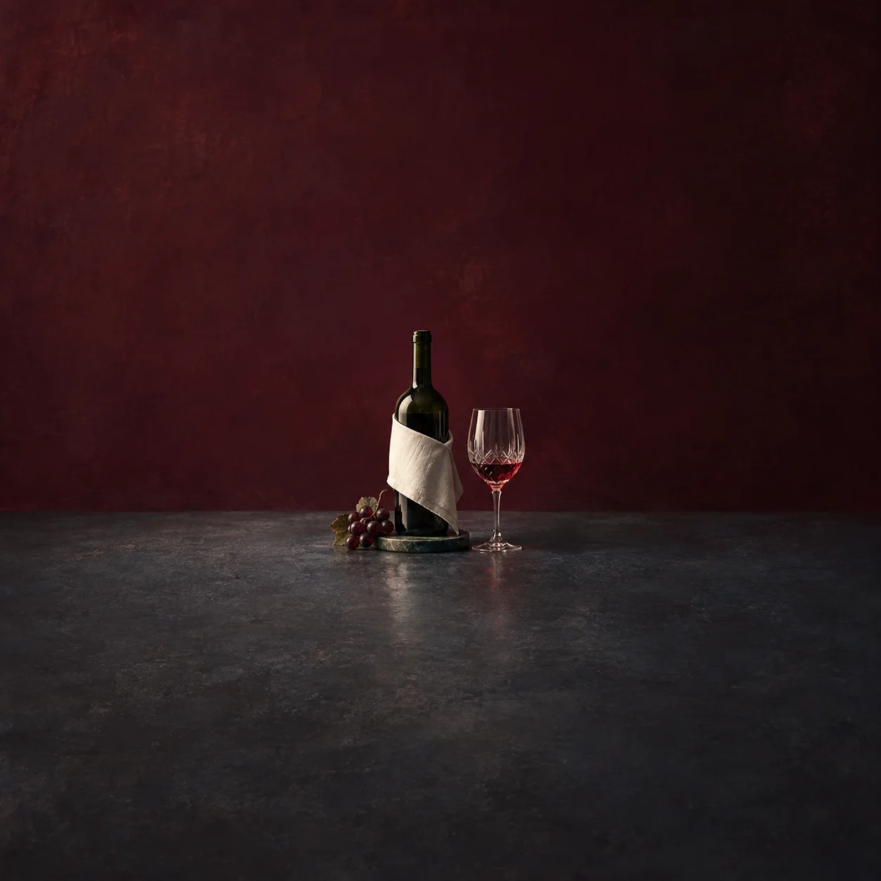 A dark green wine bottle draped with a white napkin beside a glass of red wine and a small bunch of grapes.