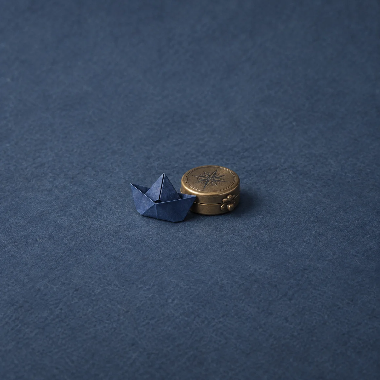 A small folded blue paper hat rests beside a closed brass compass on blue-grey felt.