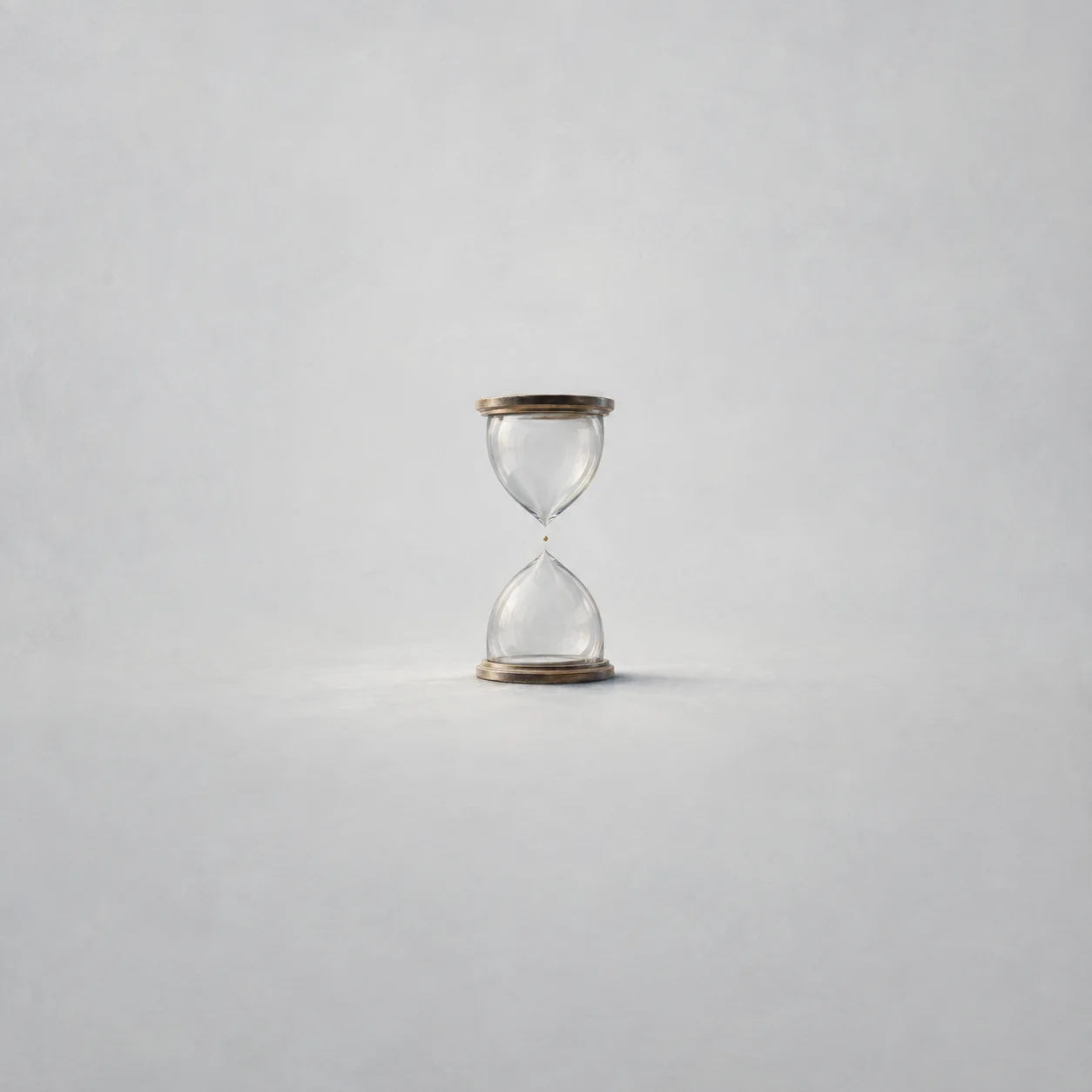 A nearly empty glass hourglass stands upright on pale grey ground with a tiny golden grain at its narrow centre.