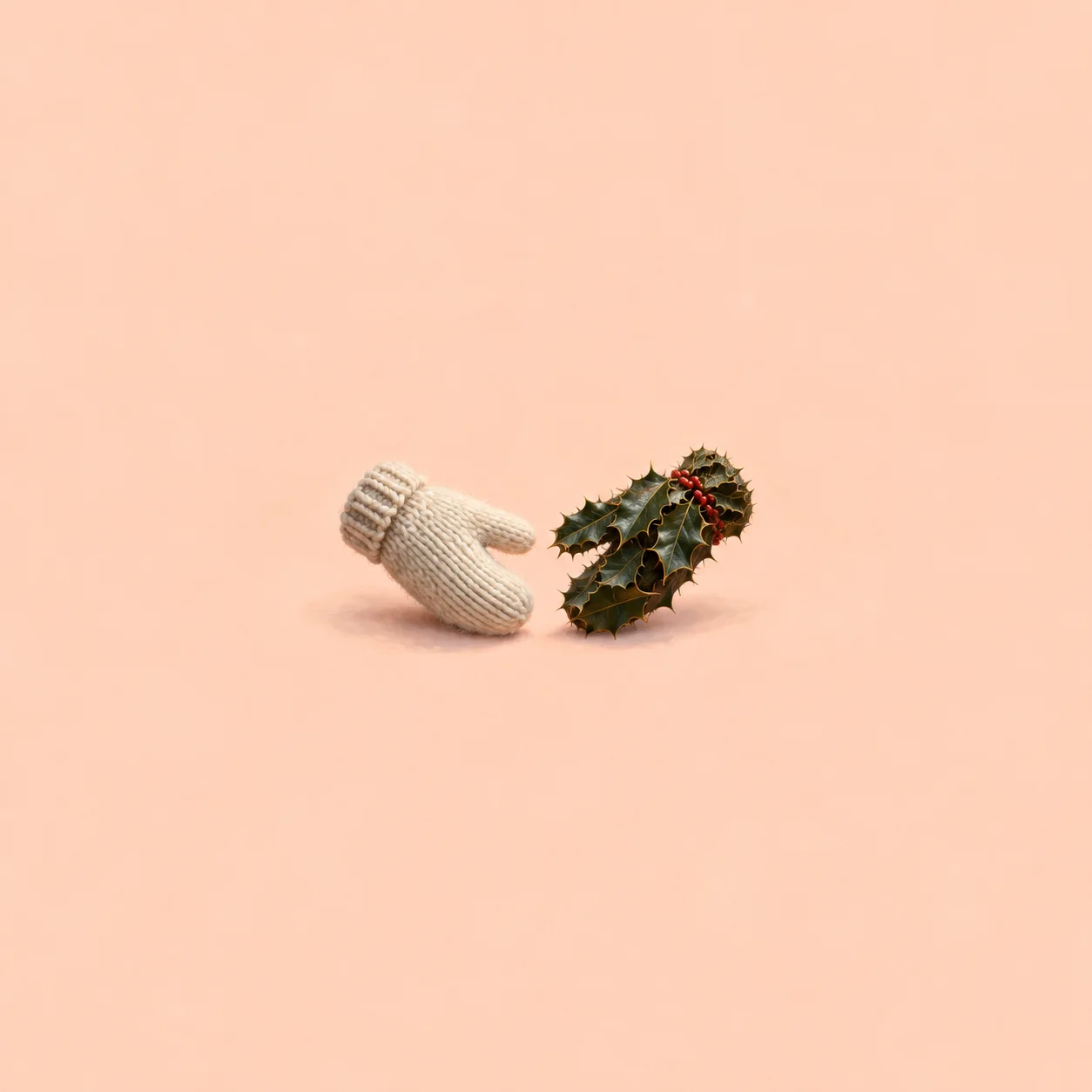 A cream knitted mitten lies beside a mitten made of holly leaves and red berries on pale peach.