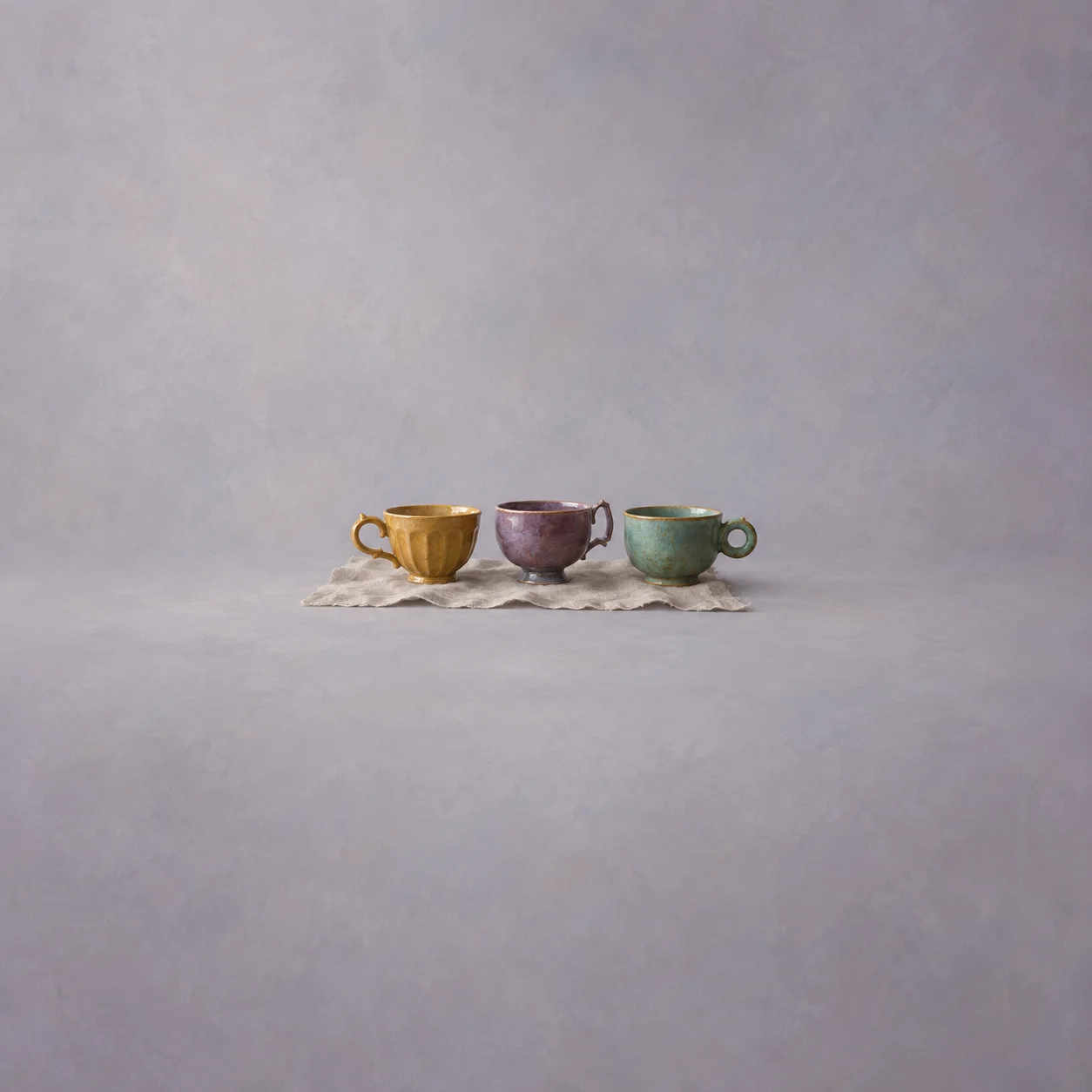 Three teacups in mustard, plum and green standing on a pale cloth against a soft gray background.