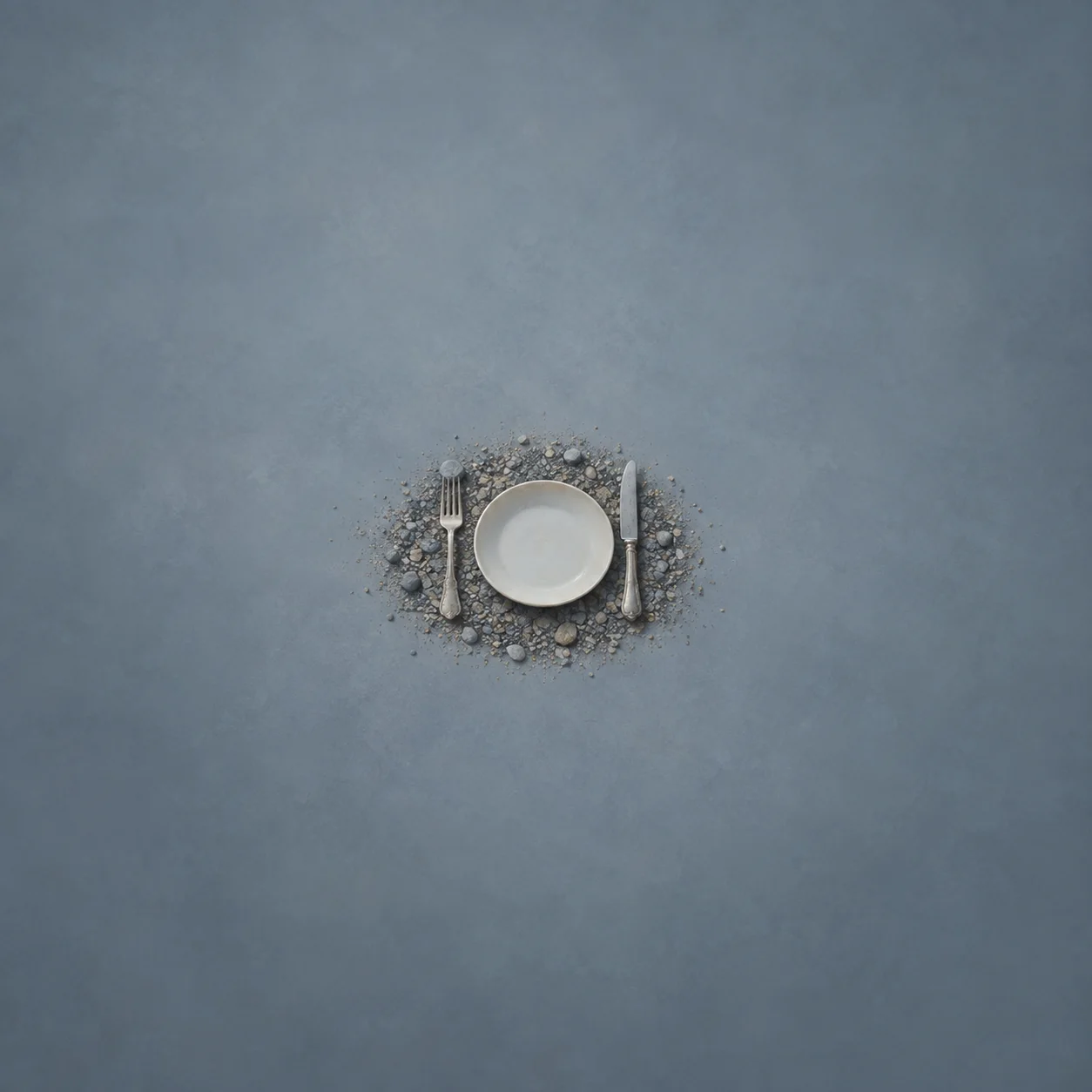 A white plate with silver cutlery rests on a small circular bed of gravel against a blue-grey background.