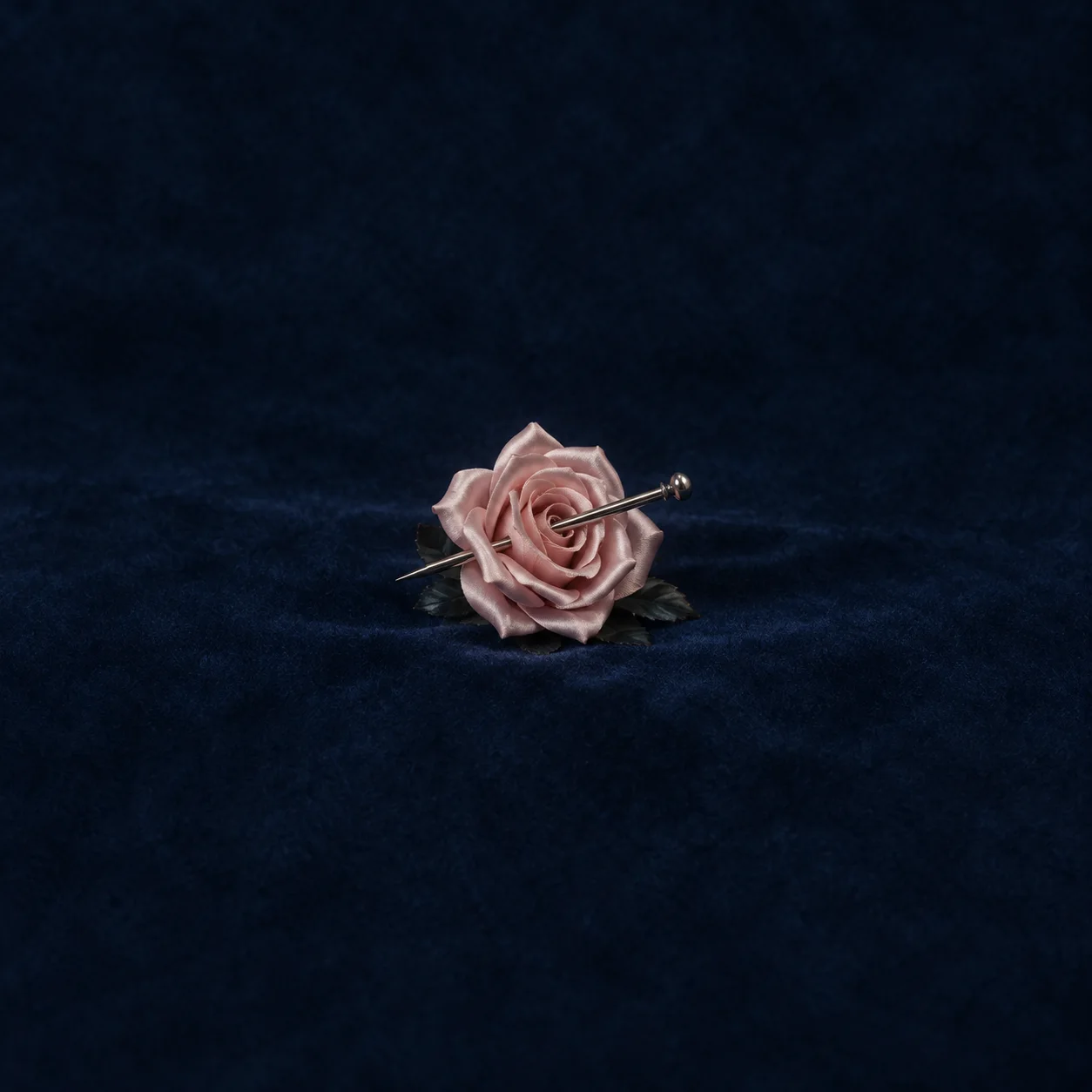 A pale pink rose pierced by a long metal pin resting on dark indigo velvet.