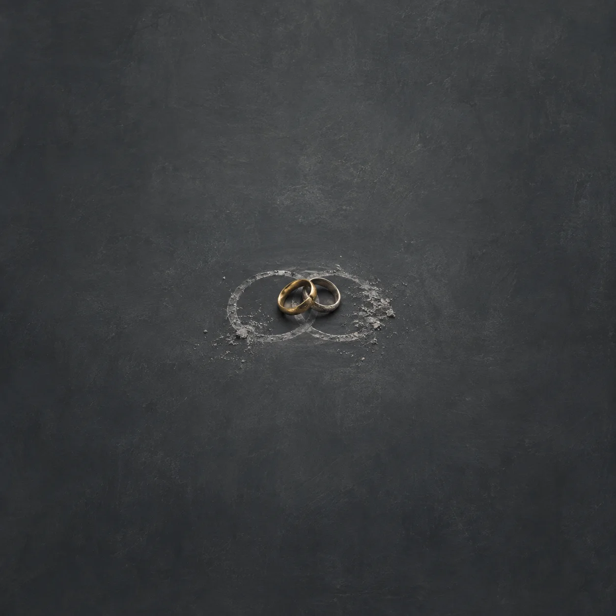 Painterly two gold wedding rings resting over overlapping chalk circles on a dark slate surface.