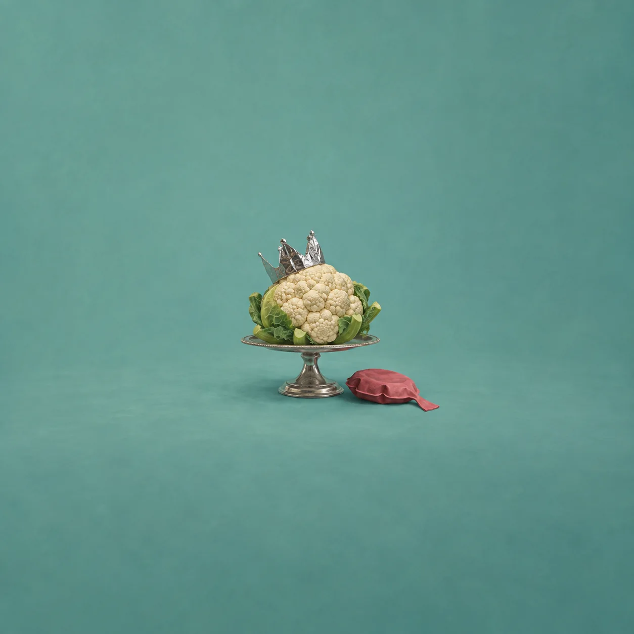 A cauliflower wearing a foil crown rests on a silver cake stand beside a pink whoopee cushion.