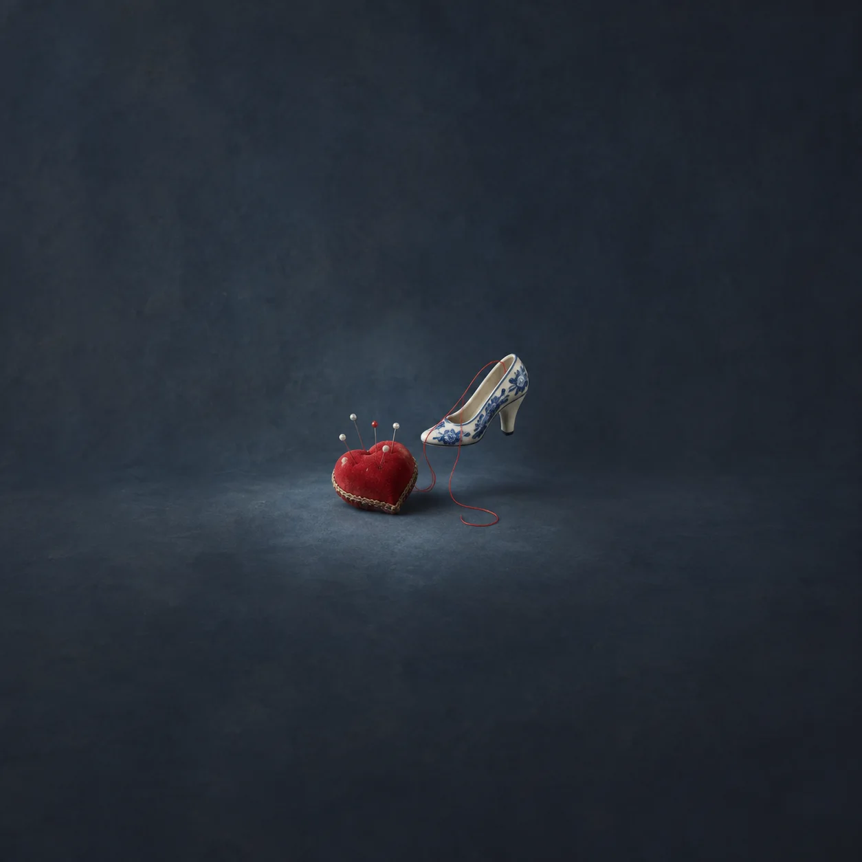 A red heart-shaped pincushion beside a hovering blue-and-white high-heeled shoe with a trailing red thread.