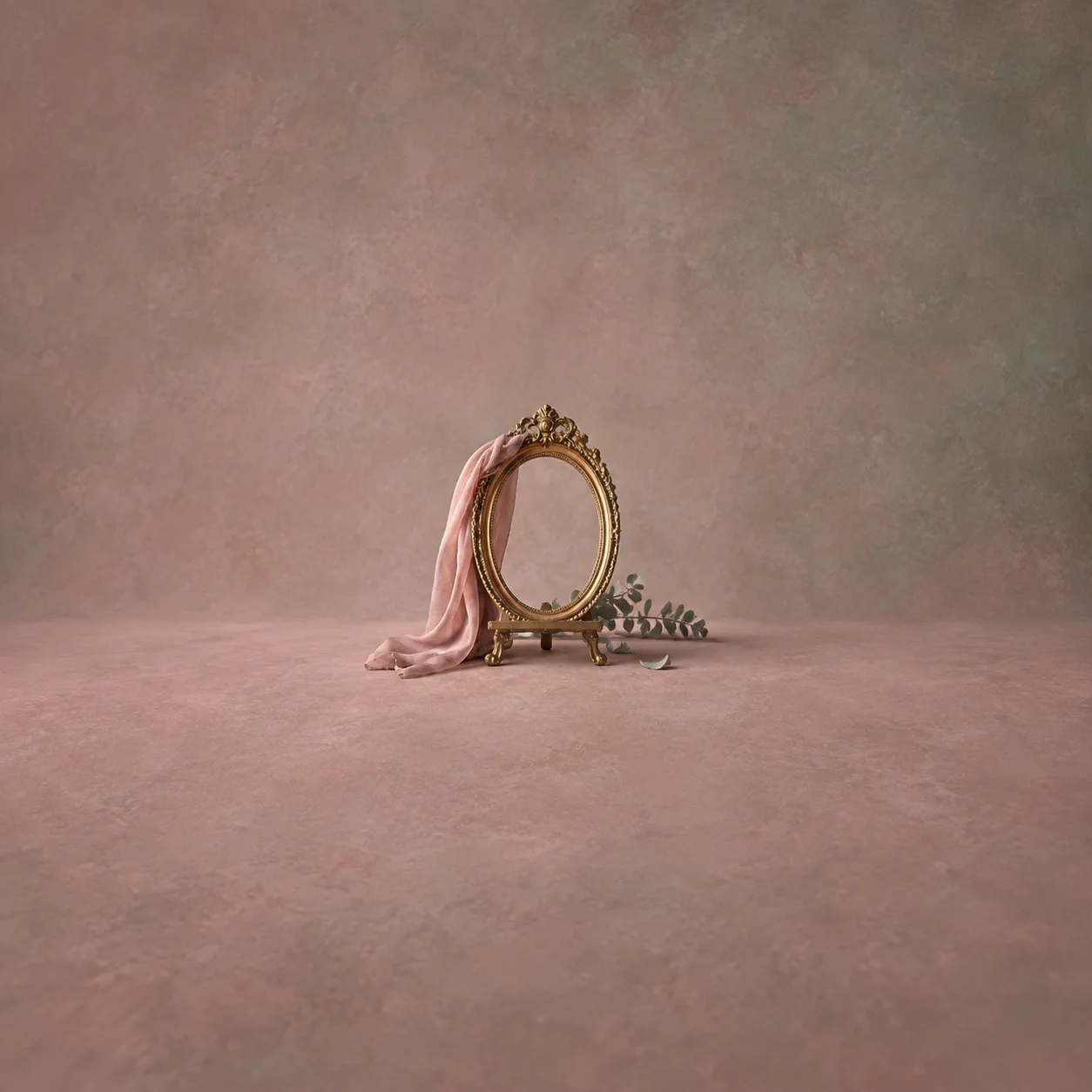 An empty oval gilt frame with pink fabric draped over one side and a leafy sprig below on a dusty-rose surface.