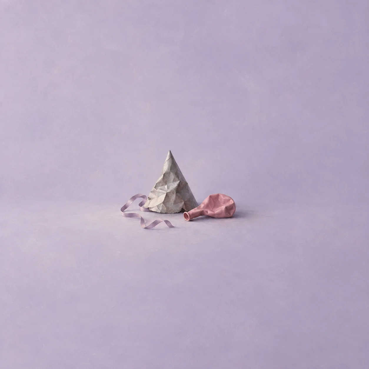 A crumpled grey party hat beside a deflated pink balloon and a loose ribbon on a lilac background.