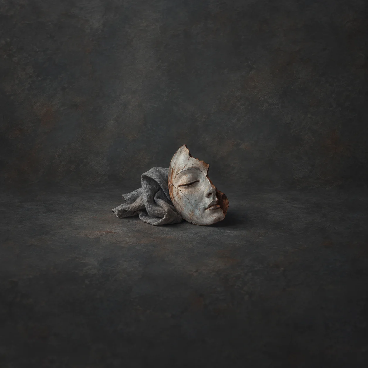A cracked pale ceramic face fragment rests beside rumpled grey cloth on a charcoal surface.