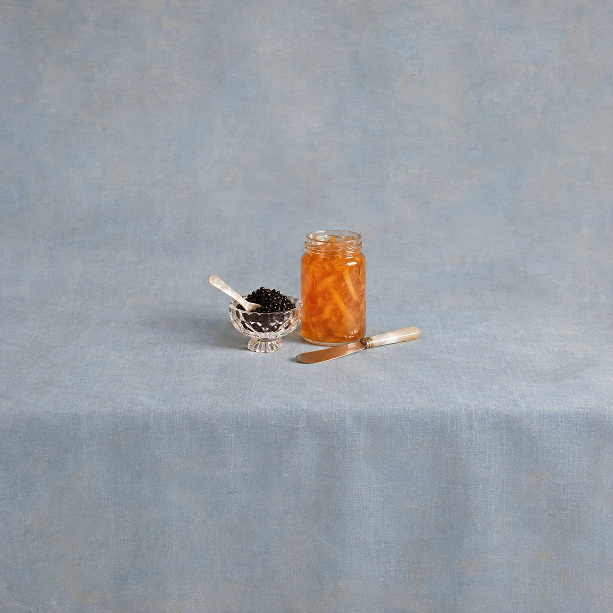 A small glass dish of black beads with a spoon beside a jar of orange marmalade and a knife.
