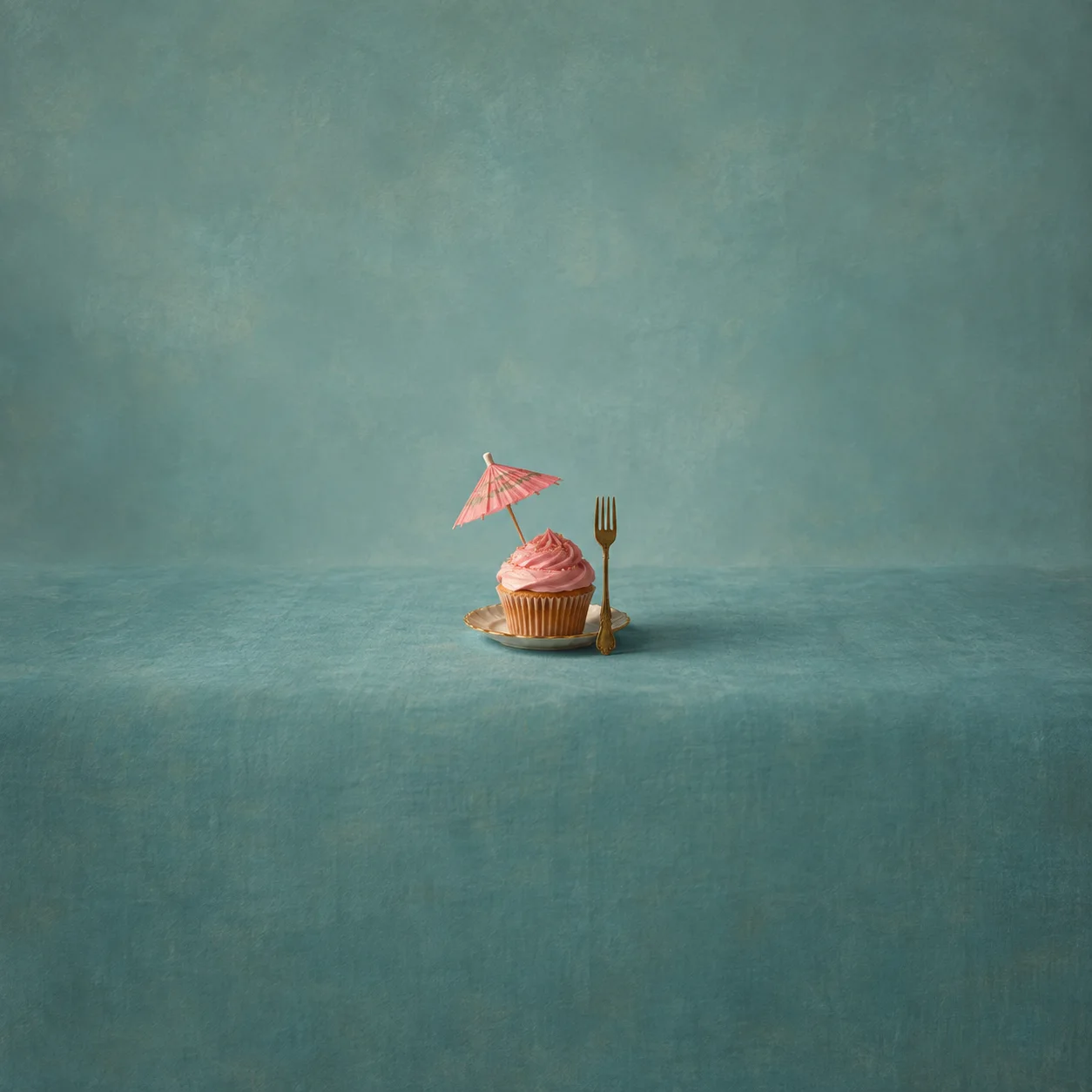 A pink-frosted cupcake with a paper umbrella sits on a small plate beside an upright gold-coloured fork on a teal surface.