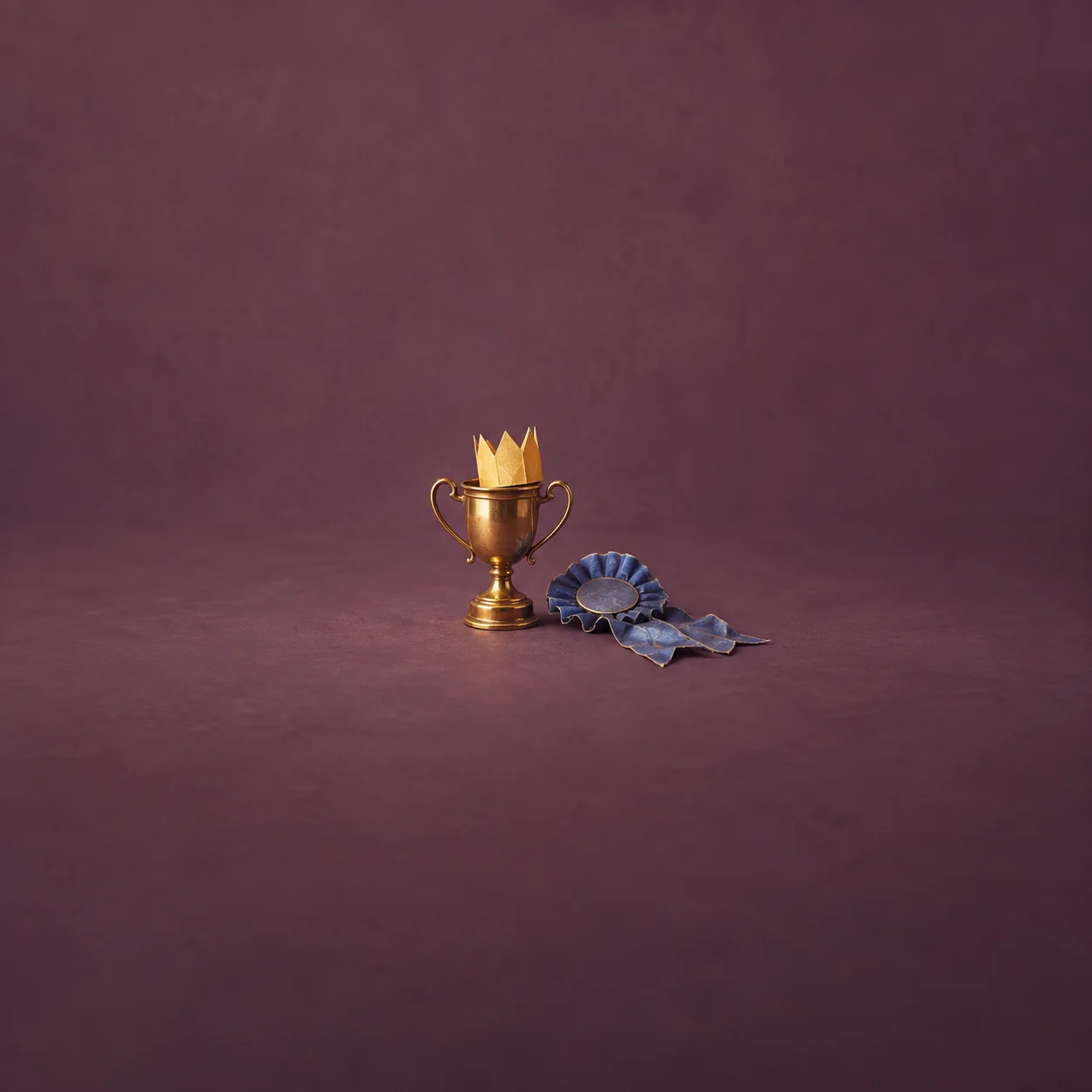 A gold trophy holds a small paper crown beside a crumpled blue rosette against purple.