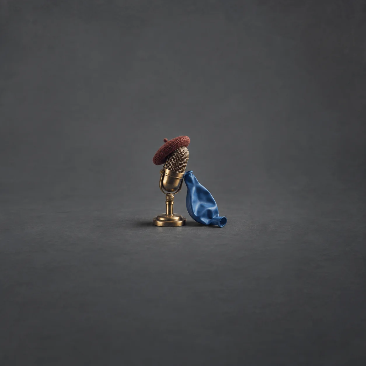 A small brass microphone wearing a tiny knitted beret with a deflated blue balloon draped beside it on a grey surface.