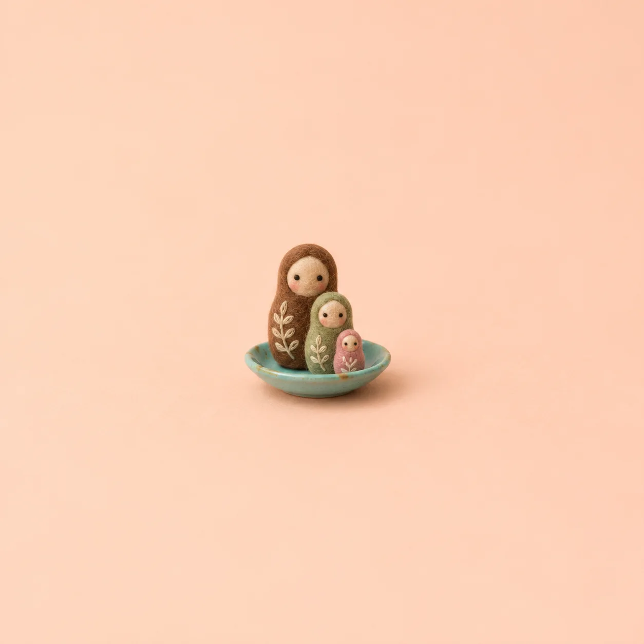 Three felt nesting figures in brown, sage and pink sit together on a turquoise saucer against peach.