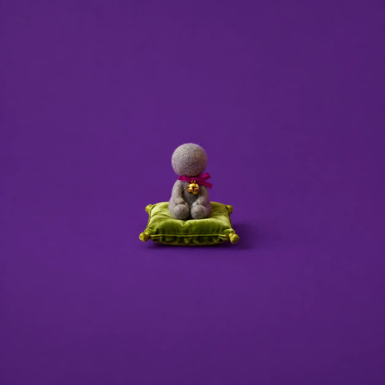 A small faceless grey felt figure wears a brass bell on magenta ribbon while sitting on a lime-green cushion.