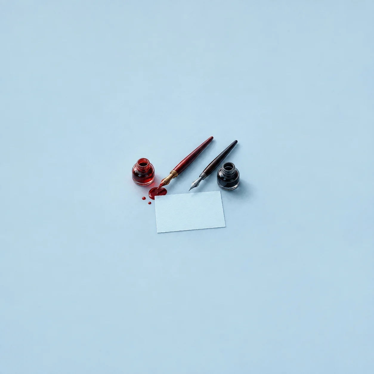 Two dip pens beside red and black ink bottles above a blank pale-blue card, with drops of red ink nearby.