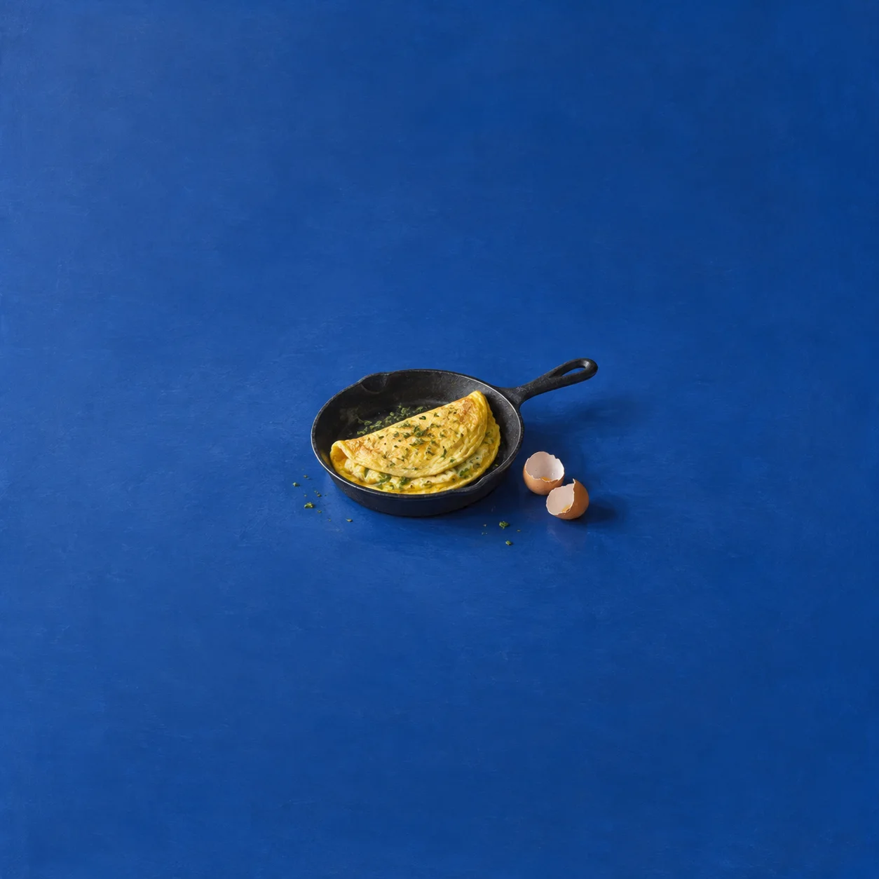 A folded omelette in a black cast-iron skillet beside two eggshell halves on a blue surface.