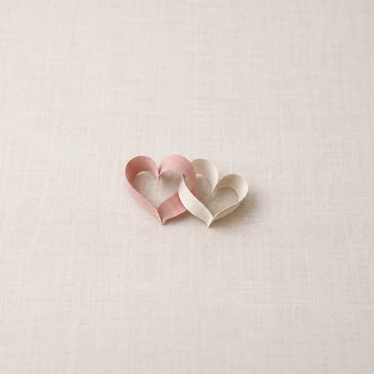 Two paper hearts, one blush pink and one cream, interlock on an ivory linen surface.