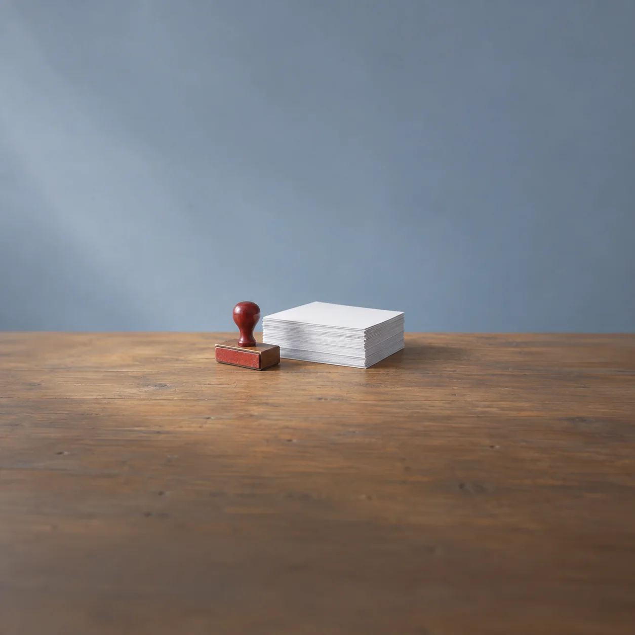 Painterly red-handled rubber stamp beside a neat stack of blank white paper on a wooden desk.