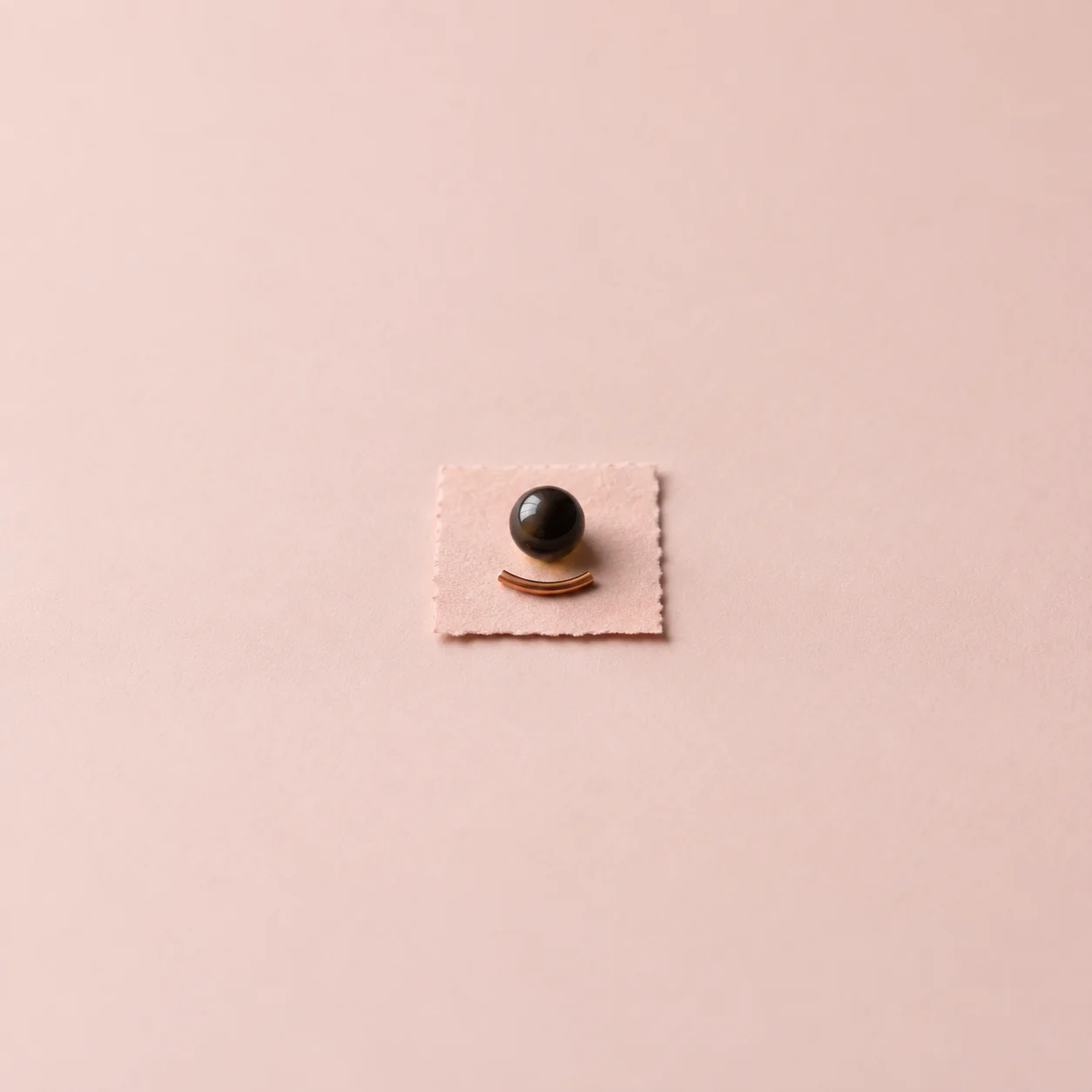 A round black bead and a short curved copper piece rest on a pale pink square.