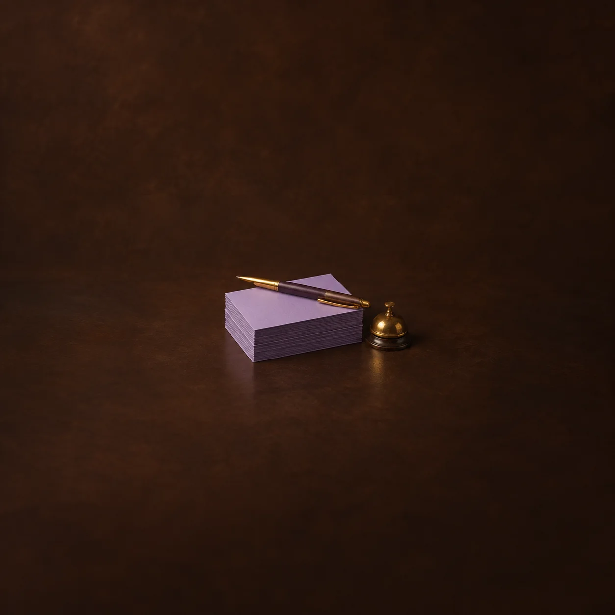 A stack of blank lilac cards with a gold-tipped pen and a small brass bell on a brown surface.