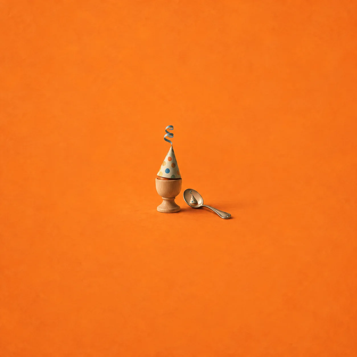 A cardboard party hat on a wooden egg cup with its reflection in a small spoon on an orange surface.