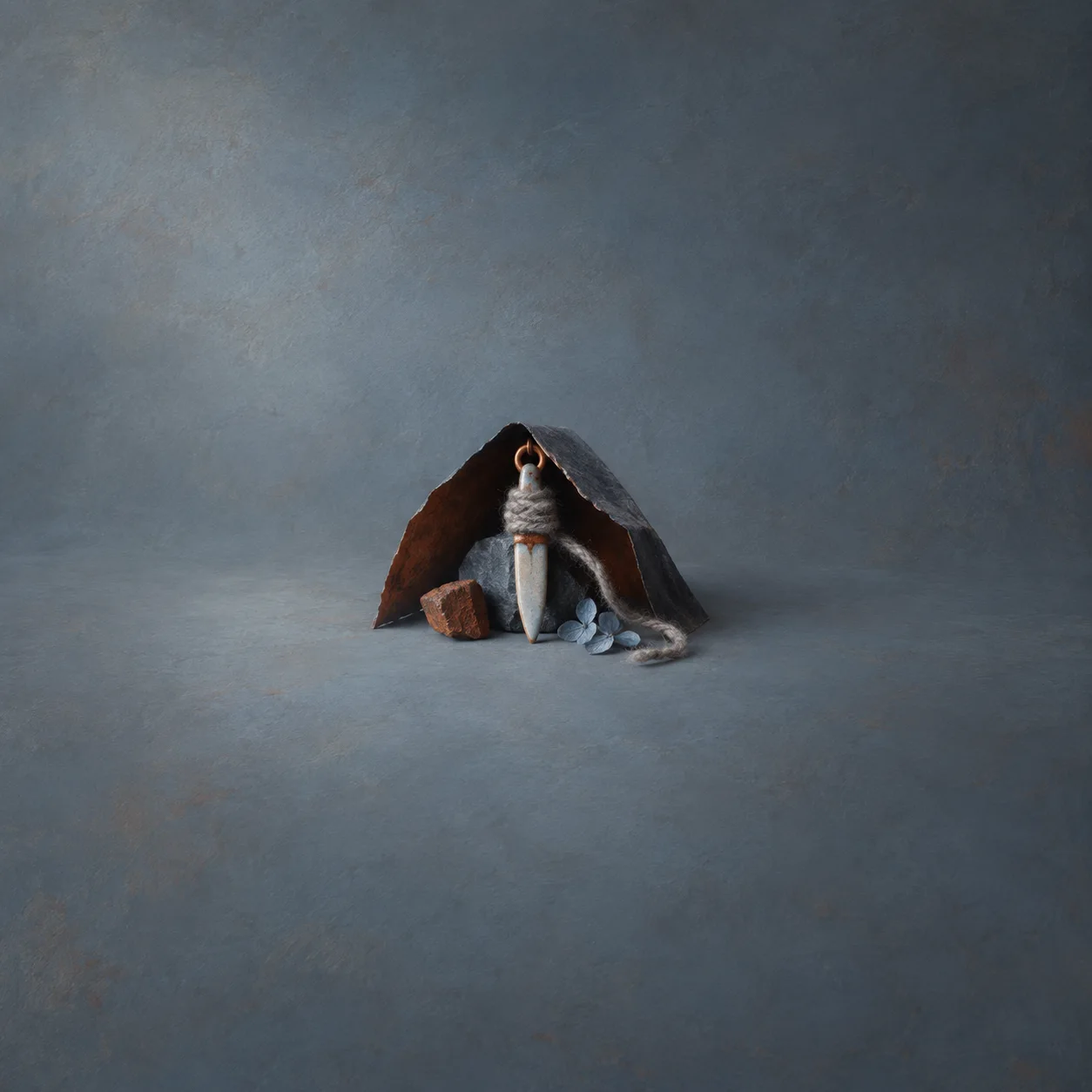A small pale dagger-shaped charm wrapped in grey thread stands beneath a dark folded arch beside pebbles.