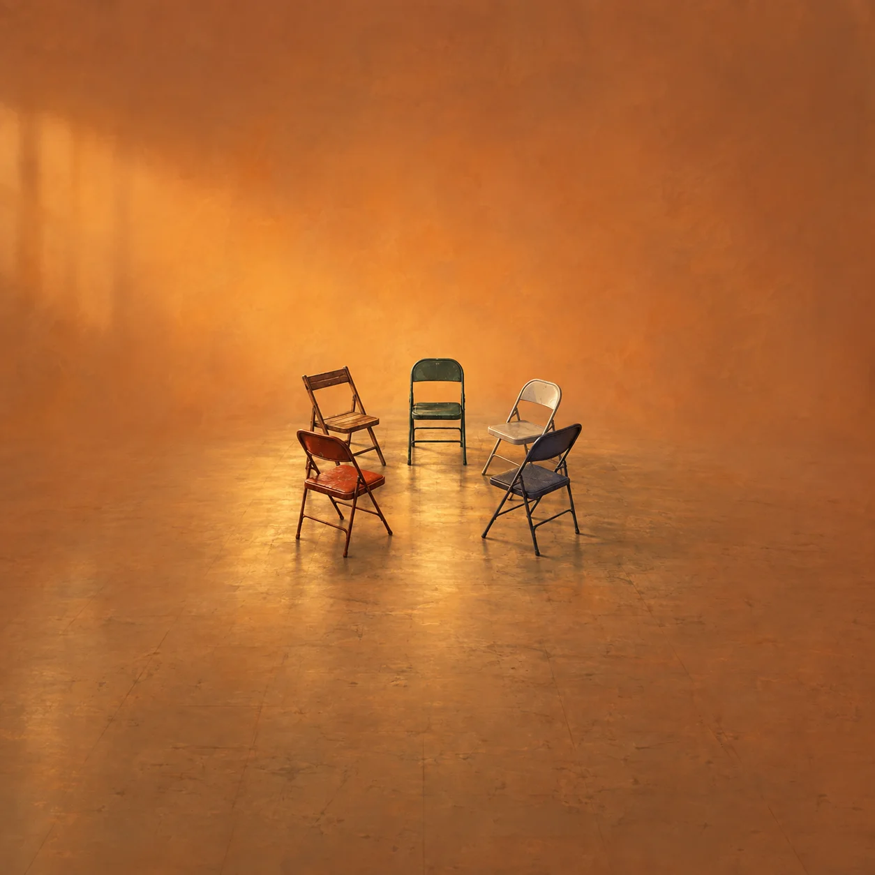 Five mismatched folding chairs form a loose circle on a scuffed floor in warm orange light.
