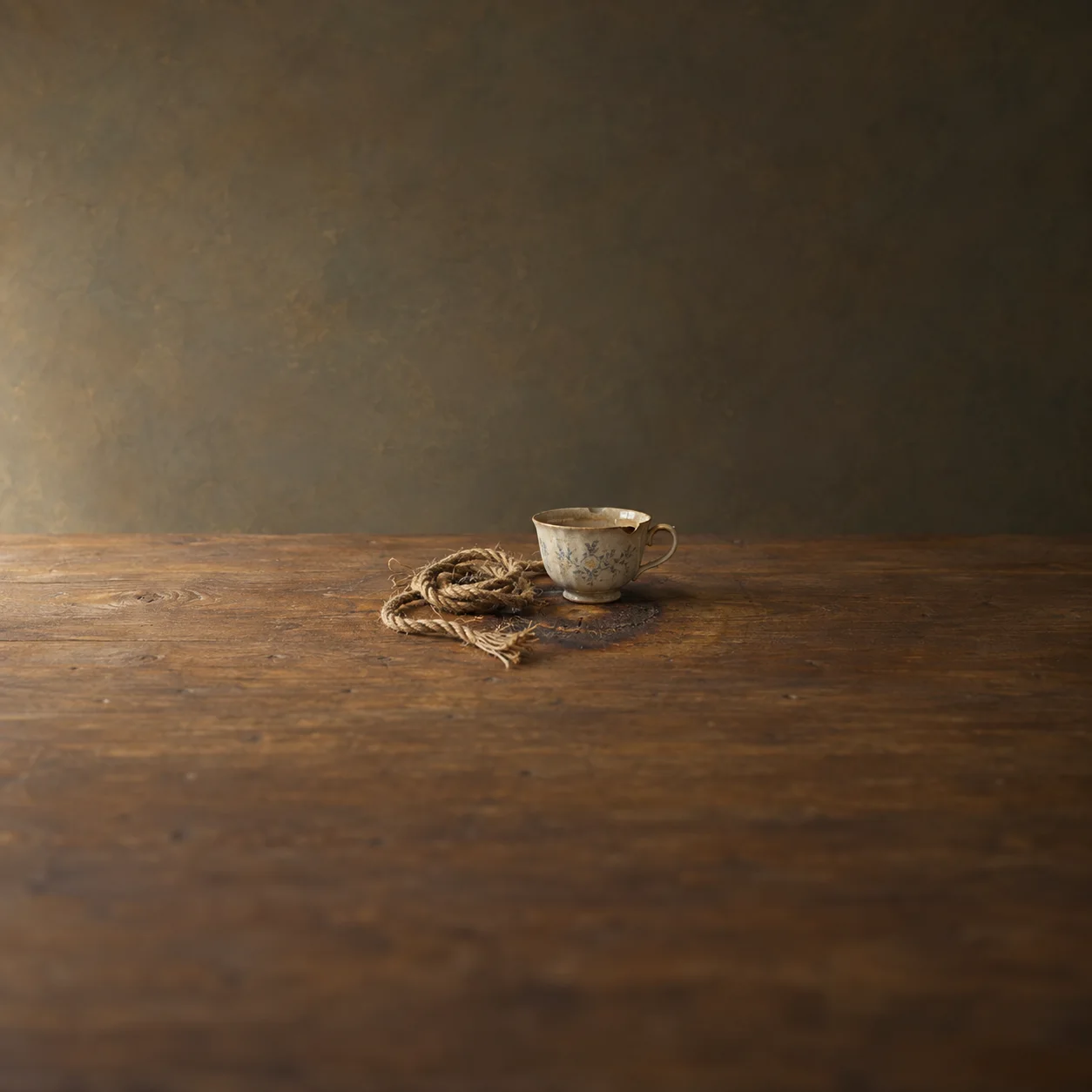Painterly coil of frayed rope beside a chipped teacup on a worn wooden table.