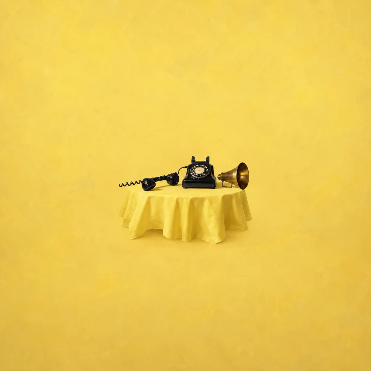 A black rotary telephone with a stretched coiled cord and a small brass megaphone beside it on yellow cloth.