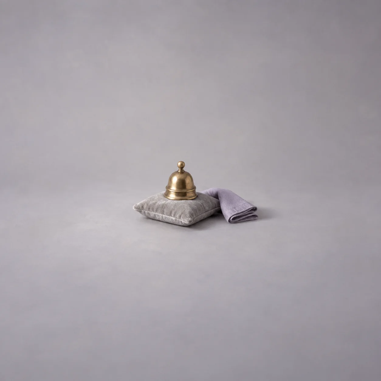 A brass bell on a grey cushion beside a folded lilac napkin.