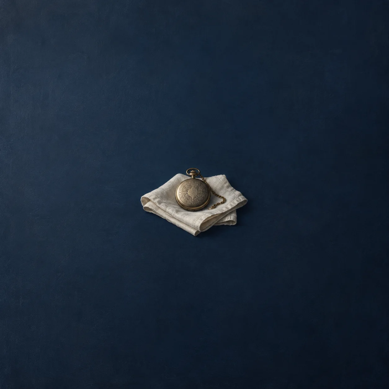 A closed metal pocket watch rests on folded white linen against dark navy.