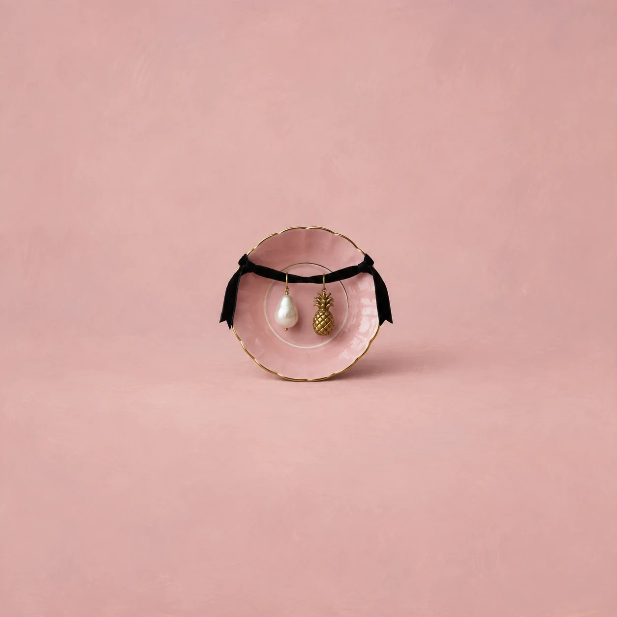 A pearl earring and a small brass pineapple earring hanging from a black ribbon above a pink saucer.