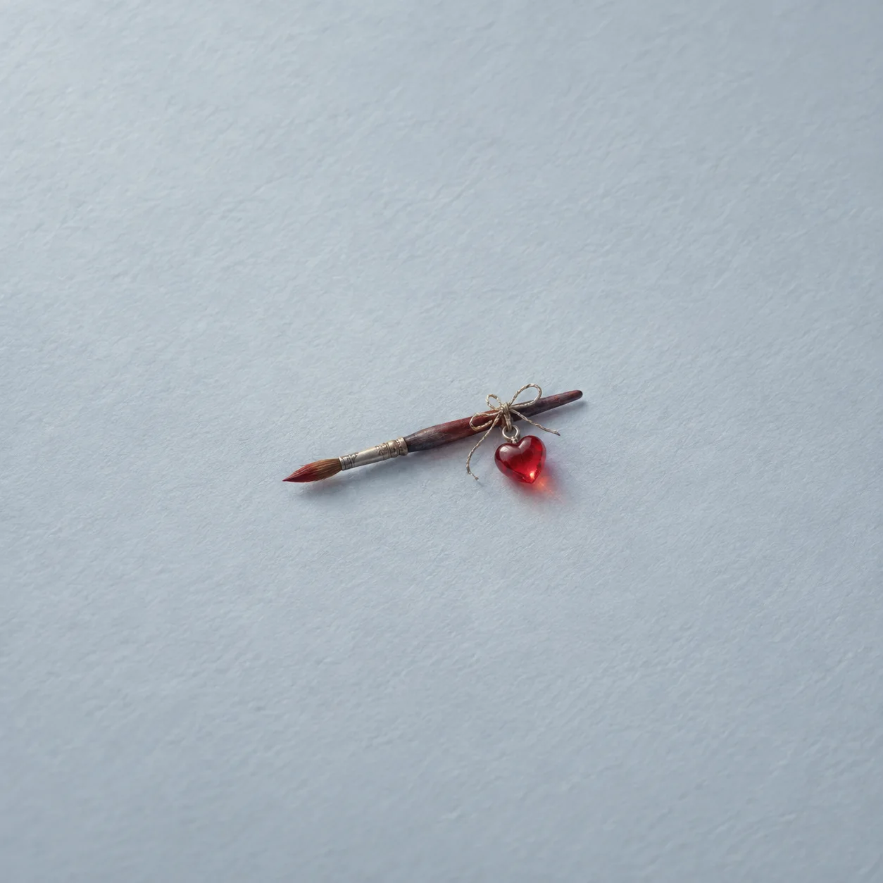 A paintbrush with a small red glass heart tied to its handle on grey-blue paper.