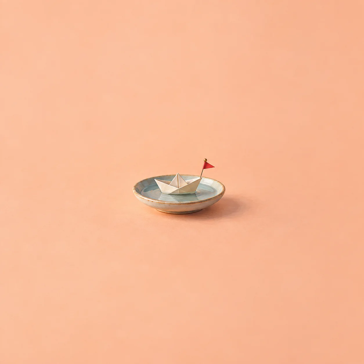 A small paper boat on a saucer of water with a tiny pushpin flag on a peach surface.