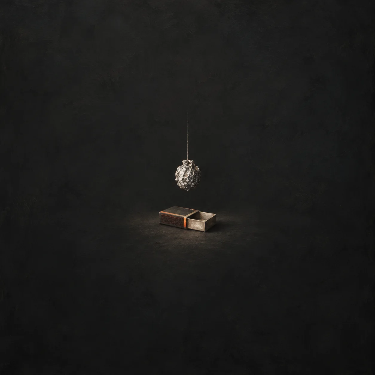 A crumpled foil ball hanging on a thread over an empty matchbox on a charcoal background.