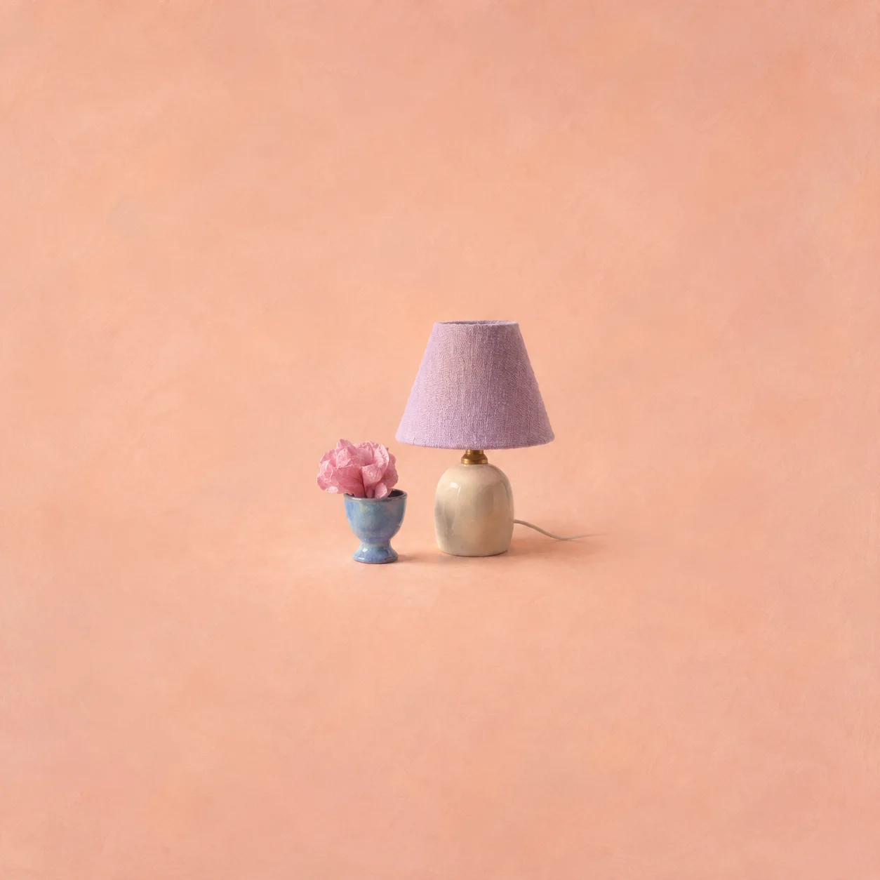 A crumpled pink tissue flower in a blue eggcup beside a small lamp with a lilac shade.
