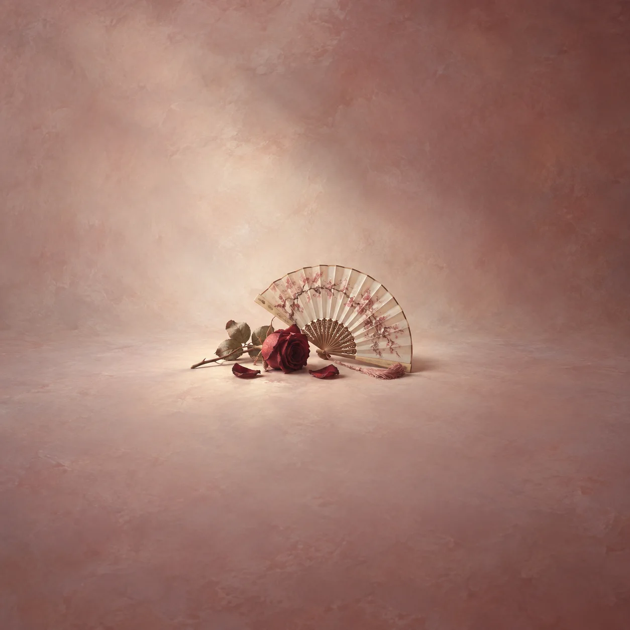 Painterly cream folding fan beside a red rose and fallen petals on dusky pink.