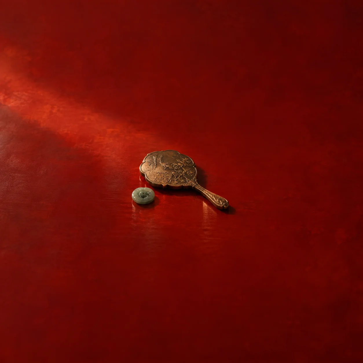 Painterly ornate hand mirror lying face-down beside a green button on red lacquer.