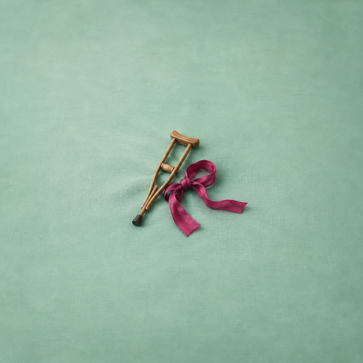 A small wooden crutch lies beside a large loose magenta ribbon against mint.